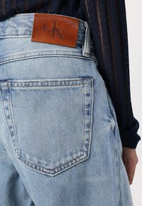 Bolsillo trasero de jeans de mezclilla azul claro con parche de cuero marrón con el logotipo de Calvin Klein, combinado con una camiseta de manga larga texturizada azul oscuro.