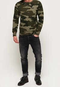 Kamerový sweatshirt v zelených odstínech s kulatým výstřihem, spárovaný s tmavě šedými džíny a černými botami. Má dlouhé rukávy a žebrované manžety.