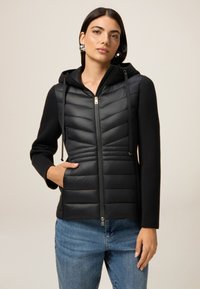 Junge Frau in einer schwarzen gesteppten Kapuzenjacke mit Reißverschluss und Jeans, die mit einer Hand in der Tasche vor einem neutralen Hintergrund posiert.
