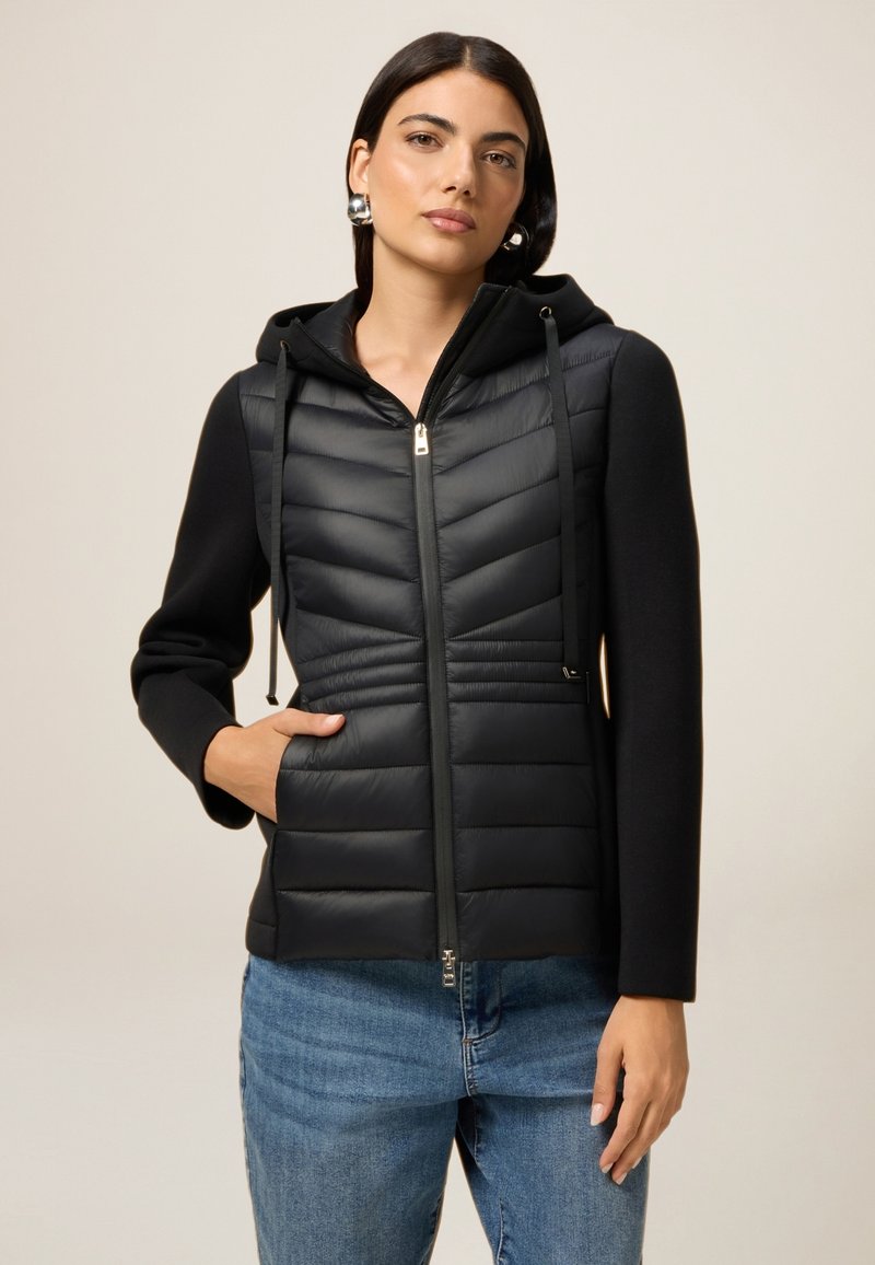 Junge Frau in einer schwarzen gesteppten Kapuzenjacke mit Reißverschluss und Jeans, die mit einer Hand in der Tasche vor einem neutralen Hintergrund posiert.