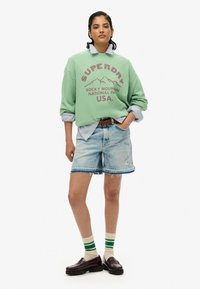 Groene sweatshirt met "SUPERDRY" logo en bergafbeelding, over een blauw gestreept shirt. Licht denim shorts, bruine loafers en gestreepte sokken.