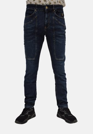 Jeans in denim scuro con vestibilità slim, dotati di tasche frontali curve, rinforzi ai ginocchi e dettagli di cucitura a contrasto. Abbinati a sneakers nere.