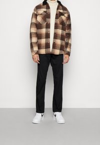 Camicia flanella a quadri marrone e beige con colletto nero, abbinata a pantaloni neri e sneakers bianche con dettagli neri.