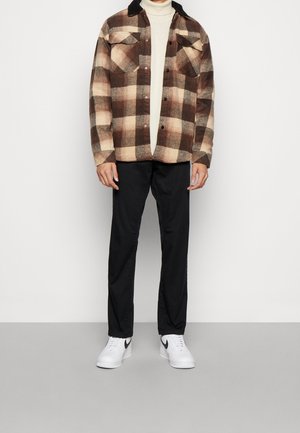 Chemise en flanelle à carreaux brun et beige avec un col noir, associée à un pantalon noir et des baskets blanches avec des accents noirs.