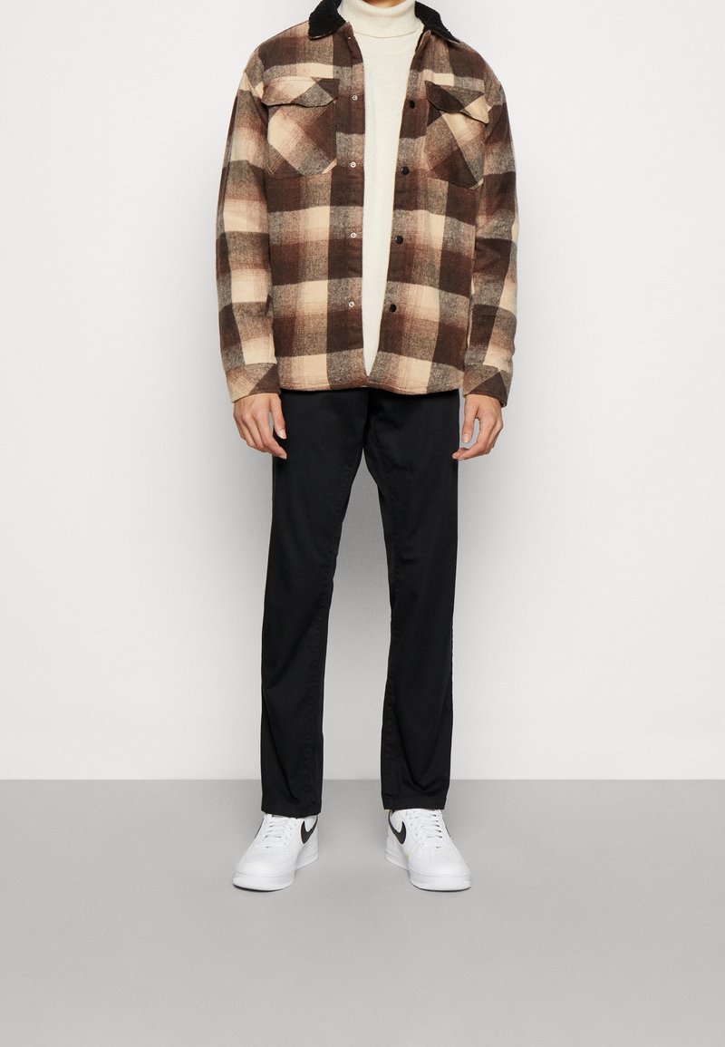 Camicia flanella a quadri marrone e beige con colletto nero, abbinata a pantaloni neri e sneakers bianche con dettagli neri.