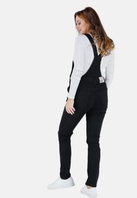 Zwarte denim overalls met een aansluitende pasvorm, gedragen over een wit lange mouwen shirt, met een logo-label en een achterzak-detail.