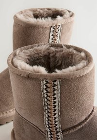 Botines de ante marrón con un suave forro de piel sintética y costuras decorativas en el costado. El diseño presenta un cuello redondeado y una superficie texturizada.
