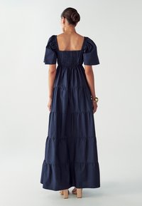 Námořnická modrá maxi šaty s nařasenými krátkými rukávy, čtveratým výstřihem na zádech a vrstvenými volány. Obsahuje nařasenou část v oblasti pasu.