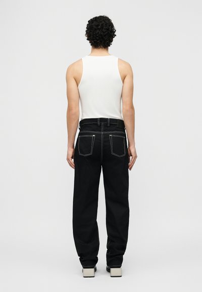 Άτομο με σγουρά μαλλιά που φοράει λευκό αμάνικο τοπ, μαύρο παντελόνι με λευκή ραφή και παπούτσια με λευκή σόλα, στέκεται με την πλάτη προς τα εμπρός.