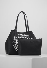 Czarna skórzana torba typu tote z białym nadrukowanym logo, w zestawie z mniejszą czarną saszetką z wytłoczonym nazwą marki. Gładka faktura, strukturalny design.