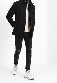 Man med svart knappad kappa, vit tröja, rivna svarta skinny jeans och vita sneakers, som står med händerna i fickorna.