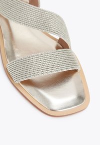 Sandal in oro metallico con suola piatta, caratterizzata da cinghie in tessuto intrecciato, design a punta quadrata e soletta in finta pelle liscia. Sottile texture e brillantezza.