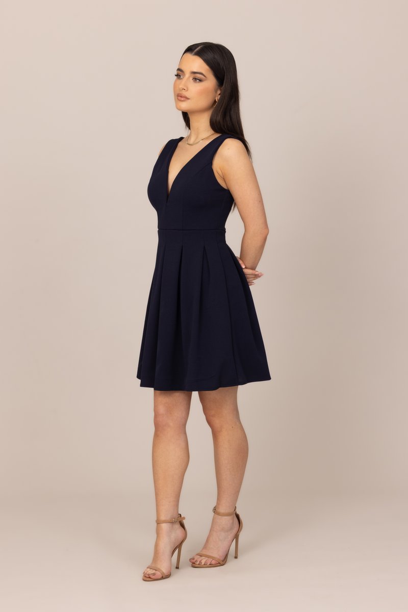 Γυναίκα με αμάνικο μπλε φόρεμα σε σχήμα V και γυμνά ψηλοτάκουνα σανδάλια, στέκεται με τα χέρια πίσω από την πλάτη, κοιτάζοντας προς το πλάι.