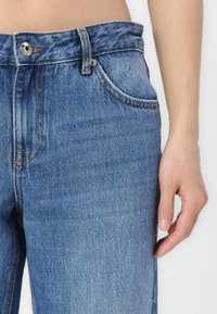 Jeans de mezclilla azul con una cintura alta, bolsillo frontal visible y costuras naranjas en contraste. La tela tiene un acabado texturizado y desvaído.