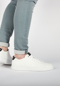Sneaker in pelle bianca con suole piatte e punte rotonde. Presentano dettagli neri sul tallone e accessori dorati per i lacci. Abbinati a jeans azzurri chiari.