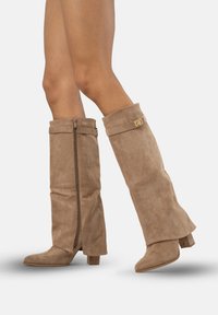 Knee-high beige mockask stövlar med klackar, dragkedjedetalj på insidan och en vikbar krage med en gyllene accent.