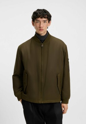 Olive green bomber jacket with a zip front, ribbed collar and cuffs, side pockets, and a zippered sleeve pocket. Features a smooth, matte finish.