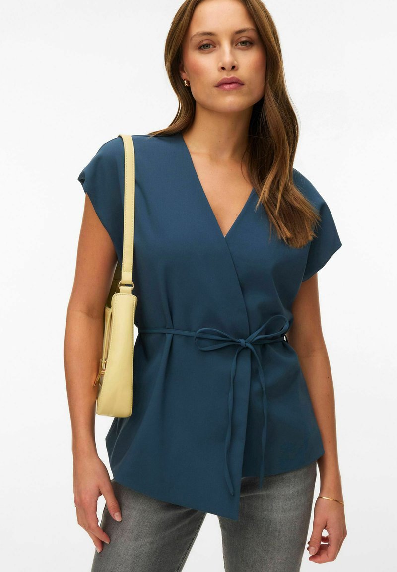 Femme portant un haut cache-cœur bleu sarcelle avec une ceinture à la taille, un jean gris, un bracelet en or, de petites boucles d'oreilles en or, et un sac à bandoulière jaune pâle.