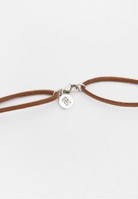 Bracciale in suede marrone con chiusura in tonalità argento e un ciondolo circolare che presenta un logo. Il design è semplice e minimalista.