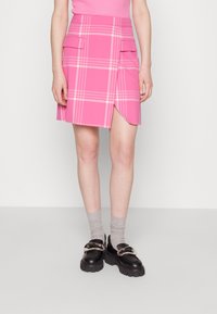 Rosa karierte A-Linien-Rock mit einem seitlichen Schlitz, ausgestattet mit zwei Fronttaschen und einer glatten Textur. Getragen mit grauen Socken und schwarzen Plateau-Schuhen.