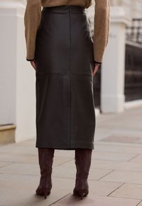 Tmavě zelená kožená midi sukně s přiléhavým střihem, svislými švy a hladkou texturou, doplněná tmavě taupe kozačkami ke kolenům.