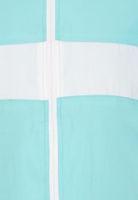 Letvægtsjakke i teal med en hvid vandret stribe. Har en central lynlås og et struktureret materiale. Renset, moderne design.