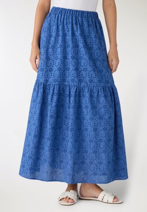 Gonna midi a balze blu con motivo a occhielli indossata da una persona con top bianco e sandali bianchi slide, in piedi su sfondo bianco.