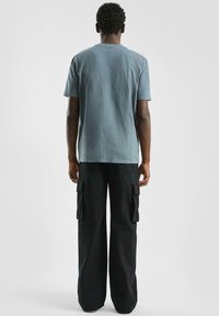 Man met korte krullende haren, gekleed in een vervaagde blauwe kortekoope T-shirt en losse zwarte cargobroek, van achteren gezien tegen een effen achtergrond.