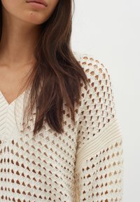 Elfenbeinfarbener Strickpullover mit einem lockeren Häkelmuster, V-Ausschnitt und gerippten Akzenten an den Ärmeln. Mit markanten Rautenlöchern.