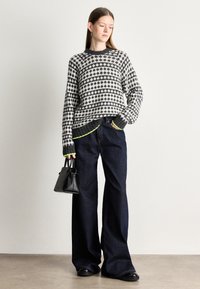 Pulover tricotat gri cu un model geometric, asociat cu blugi negri evazați. Are accente verzi neon pe manșete și o mică geantă neagră.