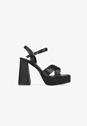 Sandalias de plataforma negras con tacón bloque. Presentan correas entrecruzadas y una correa ajustable en el tobillo. Textura de material sintético suave.