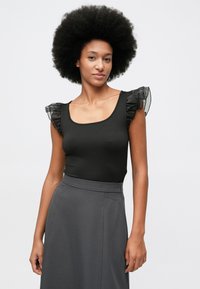 Kobieta z dużym naturalnym afro ubrana w czarny top z falbanami na rękawach i ciemnoszary spódnicą z wysokim stanem na jednolitym jasnym tle.