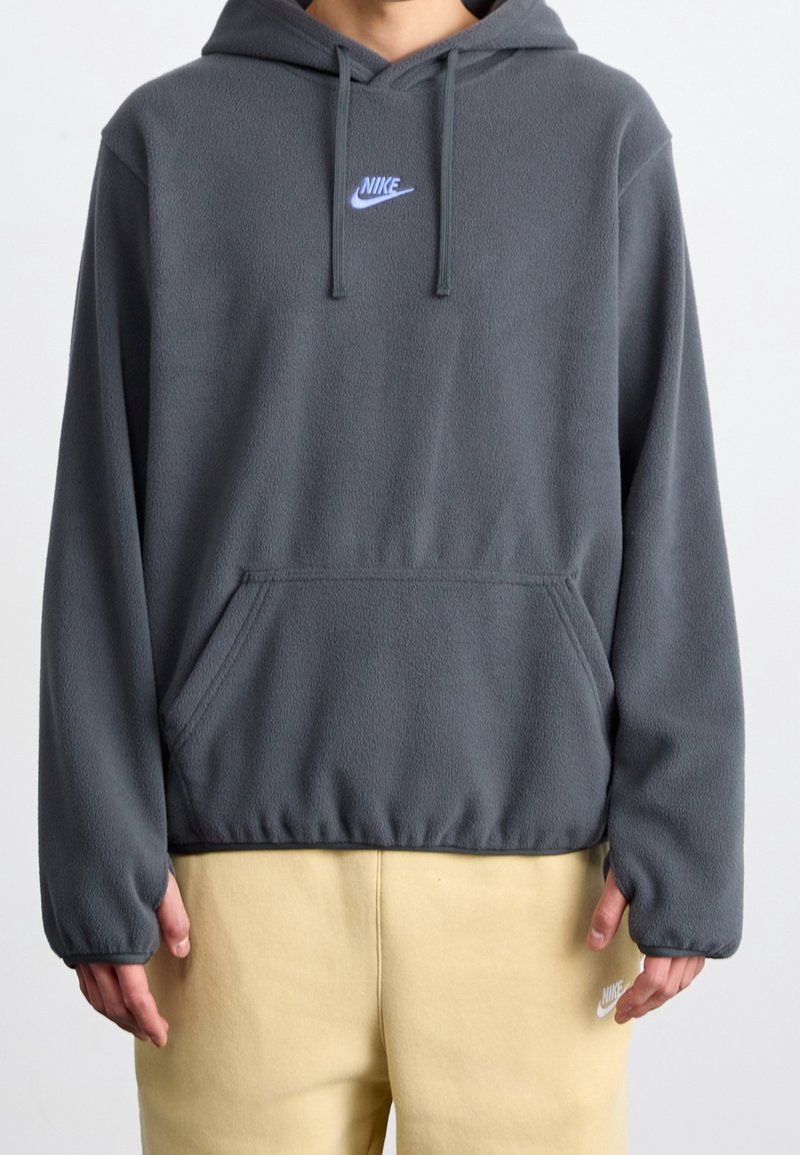 Šedá mikina s kapucí vyrobená z fleecu, s klokankovou kapsou a stahovací kapucí, na hrudi modré logo Nike. Měkká textura a volný střih.