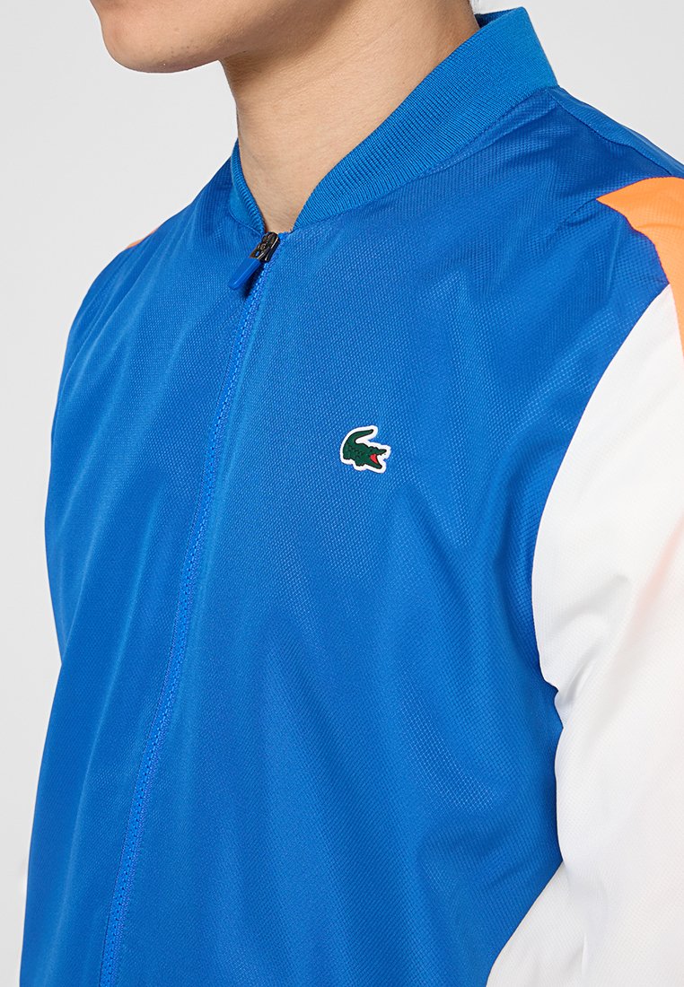 Chaqueta azul con cremallera y un tejido ligero y texturizado. Incluye un acento naranja en el hombro y un pequeño logo verde.