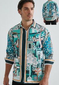 Chemise colorée à manches longues avec un motif turquoise, vert et blanc comportant des designs architecturaux et floraux. Manches longues avec des poignets contrastants.