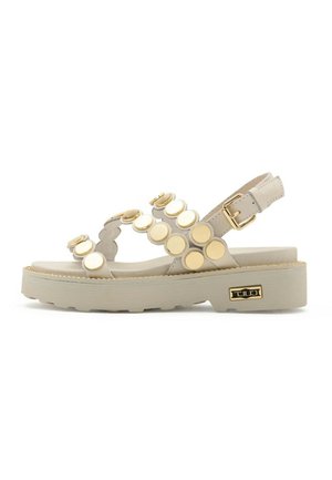 Sandalo piatto beige con suola spessa, cinturino alla caviglia regolabile e borchie decorative rotonde beige e dorate lungo i cinturini.