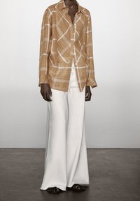 Beige geruit overhemd met lange mouwen, een kraag en knopen, gecombineerd met witte wijde broek en zwarte platte sandalen op een neutrale vloer.