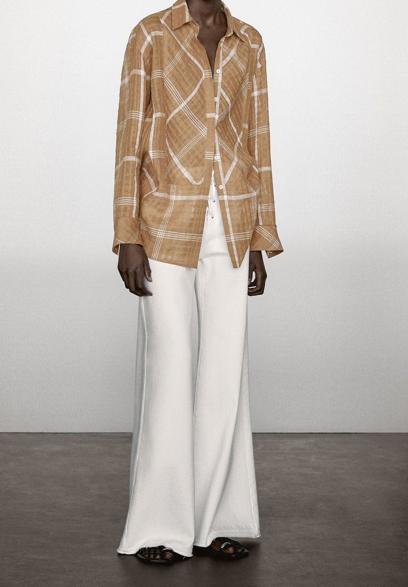 Beige geruit overhemd met lange mouwen, een kraag en knopen, gecombineerd met witte wijde broek en zwarte platte sandalen op een neutrale vloer.