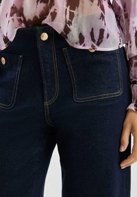 Ciemnoniebieskie dżinsy z denimu z metalowymi złotymi elementami, przednimi kieszeniami i kontrastującymi przeszyciami. Na to nałożona jest przezroczysta różowo-brązowa bluzka w wzory.