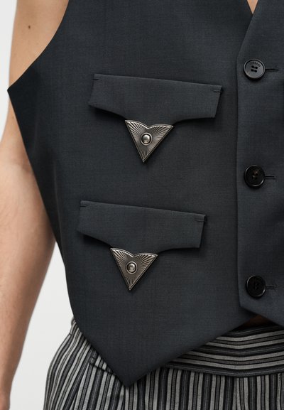 Gilet noir avec deux poches à rabat munies de boutons triangulaires argentés, porté sur un pantalon rayé.