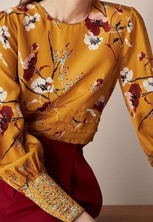 Woman wearing mustard yellow floral long-sleeve blouse with pleated cuffs and burgundy high-waisted pants against neutral background.