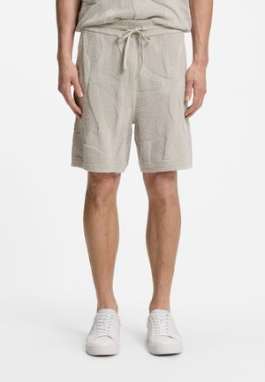 Homme portant un short beige texturé avec cordon de serrage et des baskets blanches, debout devant un fond blanc uni.