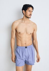 Gaiši purpursarkani kokvilnas bokseršorti ar elastīgu jostu un mazu melnu birku, ko valkā modelis ar tonizētu ķermeni.