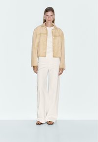 Beige suède cropped jacket met knoopsluitingen, gedragen over een witte top en gecombineerd met witte wijde broekspijpen. De outfit heeft een gladde textuur.