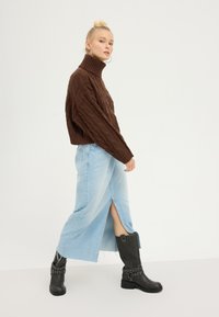 Brown cable knit turtleneck sweater paired with a light blue denim skirt featuring a front slit, and black knee-high boots with buckle accents.
