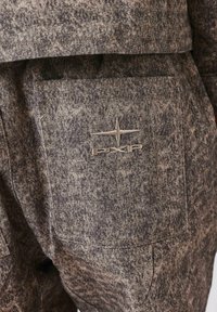 Tejido marrón texturizado con un patrón moteado, que presenta un bolsillo trasero y un logotipo bordado en un tono más claro.