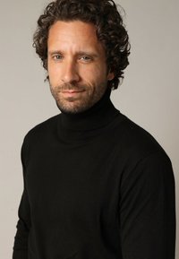 Witte man met krullend bruin haar en een baard, gekleed in een zwarte coltrui, kijkt recht in de camera met een neutrale uitdrukking.