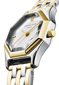 Achthochtige Uhr mit silbernem Zifferblatt, goldenen Akzenten und einem zweifarbigen Armband. Verfügt über eine markante Krone, minimalistische Indizes und ein Kristallglas.