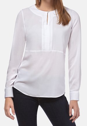Mujer con blusa blanca de manga larga con cuello redondo y pliegues verticales en la parte delantera, combinada con pantalones oscuros.