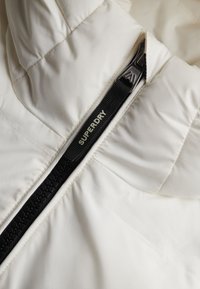 Biała kurtka puchowa o pikowanej fakturze, z czarnym suwakiem i złotym logo "SUPERDRY". Zamek błyskawiczny umieszczony centralnie.
